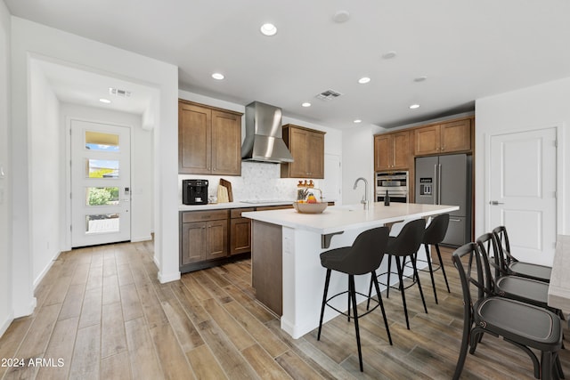 kitchen featuring a kitchen island with sink, wood-type flooring, a breakfast bar, wall chimney exhaust hood, and appliances with stainless steel finishes