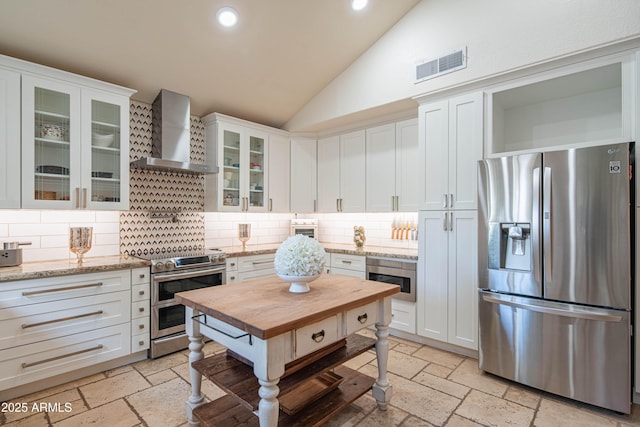 kitchen with lofted ceiling, stainless steel appliances, visible vents, wall chimney range hood, and stone tile flooring