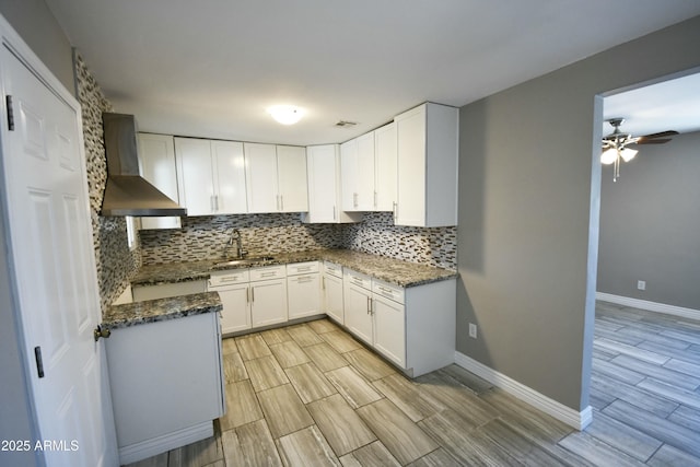 kitchen featuring wall chimney range hood, sink, dark stone countertops, tasteful backsplash, and white cabinets