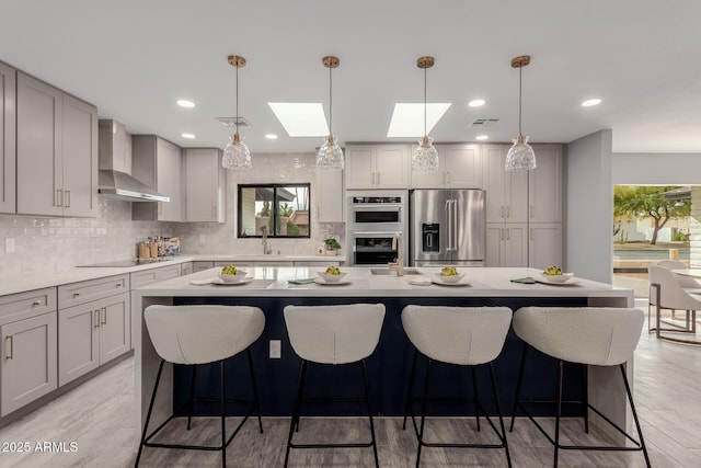 kitchen with decorative light fixtures, sink, a kitchen island with sink, stainless steel appliances, and wall chimney range hood