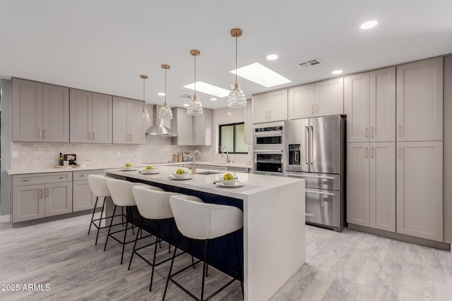 kitchen with decorative light fixtures, gray cabinets, stainless steel appliances, a kitchen island with sink, and wall chimney range hood