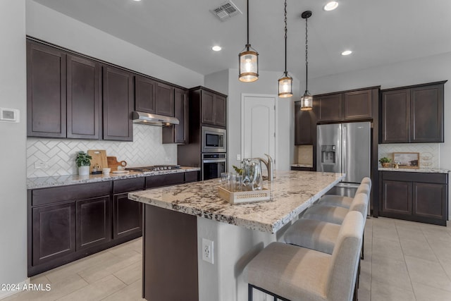 kitchen with backsplash, dark brown cabinets, stainless steel appliances, a kitchen island with sink, and decorative light fixtures