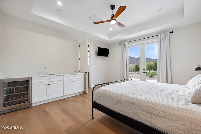 bedroom featuring beverage cooler, a tray ceiling, a sink, light wood-style floors, and access to outside