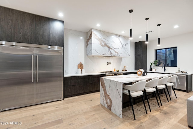 kitchen featuring decorative light fixtures, appliances with stainless steel finishes, a center island with sink, light hardwood / wood-style floors, and light stone counters