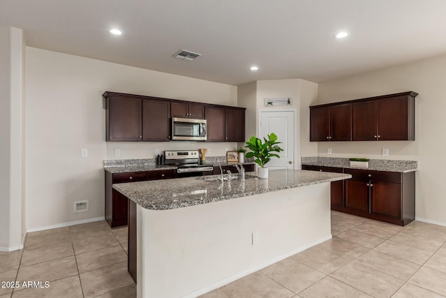 kitchen featuring dark brown cabinets, light tile patterned flooring, stone countertops, stainless steel appliances, and a center island with sink