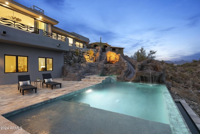 pool at dusk featuring a patio, a water slide, and a hot tub