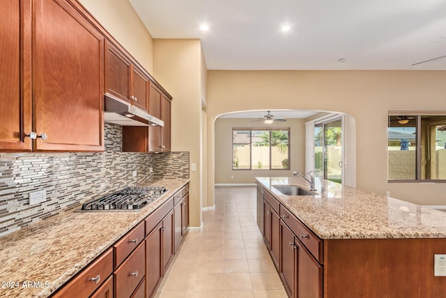 kitchen with stainless steel gas stovetop, a center island with sink, sink, ceiling fan, and light stone counters