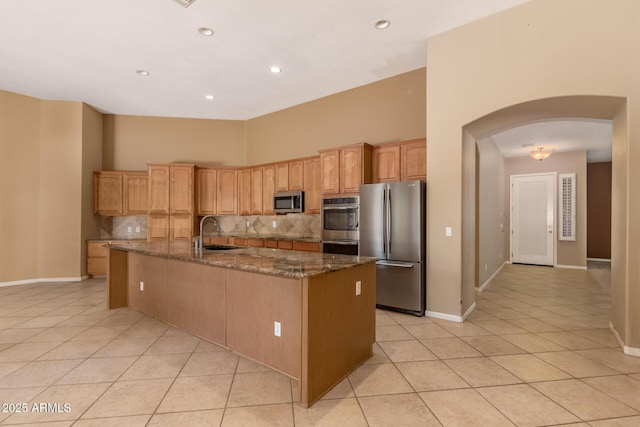 kitchen with light tile patterned floors, dark stone counters, arched walkways, a sink, and stainless steel appliances