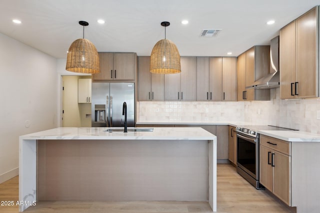 kitchen with wall chimney range hood, appliances with stainless steel finishes, a sink, a kitchen island with sink, and light wood-style floors