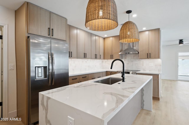 kitchen with stainless steel fridge, a sink, wall chimney range hood, light stone counters, and backsplash