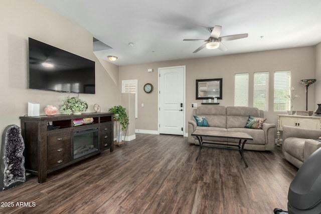 living room with dark wood-style floors, a glass covered fireplace, baseboards, and a ceiling fan