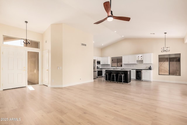 unfurnished living room with sink, light hardwood / wood-style flooring, high vaulted ceiling, ceiling fan with notable chandelier, and beverage cooler