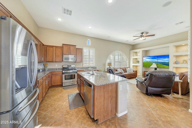 kitchen with appliances with stainless steel finishes, sink, a kitchen island with sink, light stone countertops, and built in shelves