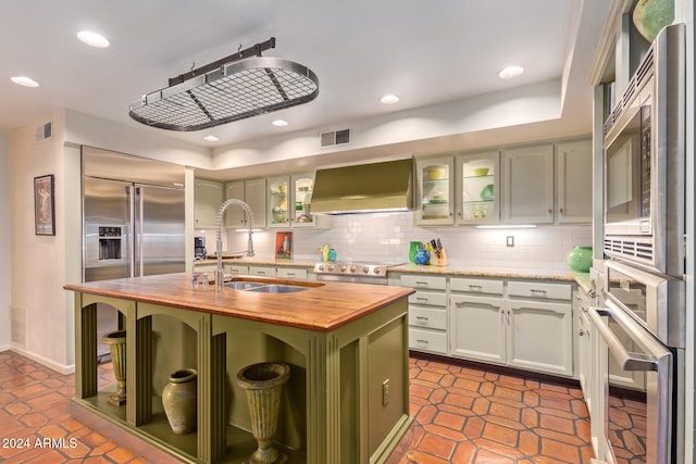 kitchen with white cabinetry, an island with sink, decorative backsplash, custom range hood, and green cabinetry