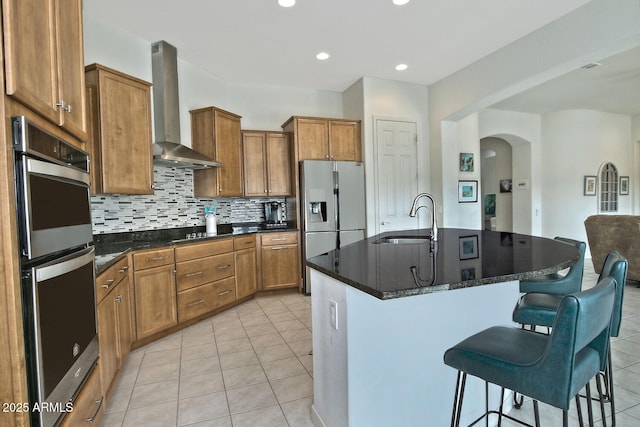 kitchen with a center island with sink, wall chimney range hood, sink, appliances with stainless steel finishes, and tasteful backsplash