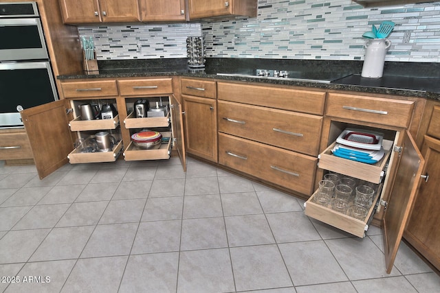 kitchen featuring stainless steel double oven, backsplash, dark stone countertops, black stovetop, and light tile patterned floors