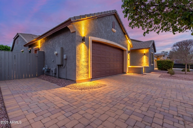 property exterior at dusk with stucco siding, an attached garage, decorative driveway, and a gate