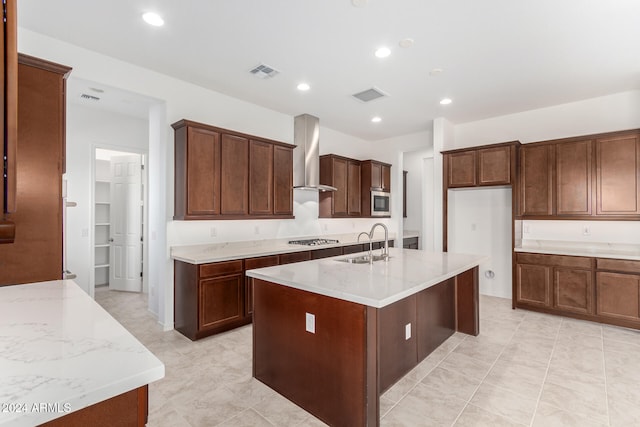 kitchen with sink, an island with sink, wall chimney range hood, light stone countertops, and appliances with stainless steel finishes