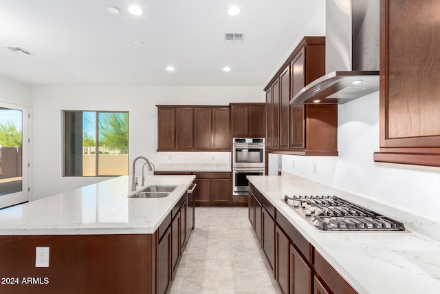 kitchen featuring light stone counters, stainless steel appliances, wall chimney range hood, sink, and a kitchen island with sink