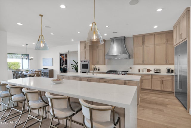 kitchen featuring pendant lighting, a large island with sink, stainless steel appliances, light brown cabinets, and wall chimney exhaust hood