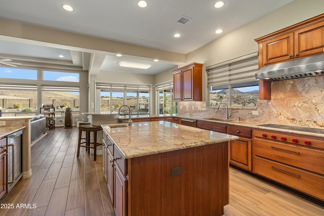 kitchen featuring an island with sink, ventilation hood, a kitchen bar, decorative backsplash, and sink