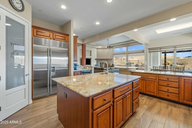 kitchen with a kitchen island with sink, built in refrigerator, light stone countertops, ceiling fan, and backsplash