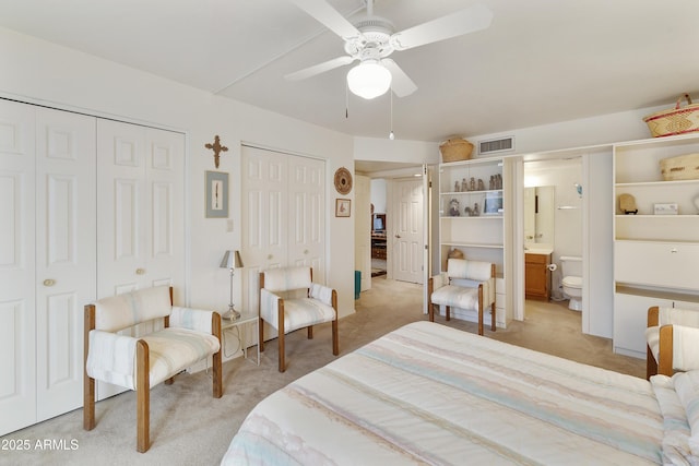 bedroom featuring visible vents, a ceiling fan, light colored carpet, ensuite bath, and multiple closets