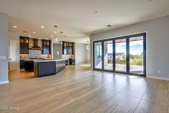 kitchen with sink, wall chimney exhaust hood, a kitchen island with sink, pendant lighting, and decorative backsplash