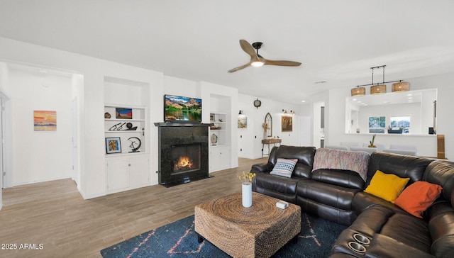 living area with built in features, a ceiling fan, a fireplace with flush hearth, and wood finished floors