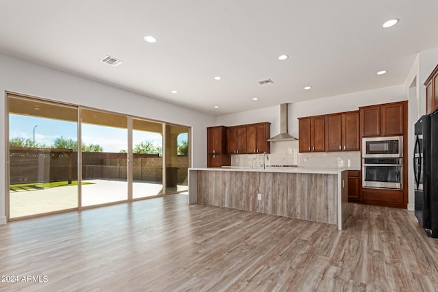 kitchen featuring appliances with stainless steel finishes, wall chimney range hood, light hardwood / wood-style flooring, and an island with sink