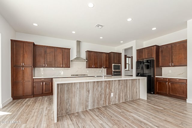 kitchen with light hardwood / wood-style flooring, wall chimney exhaust hood, a kitchen island with sink, and stainless steel appliances