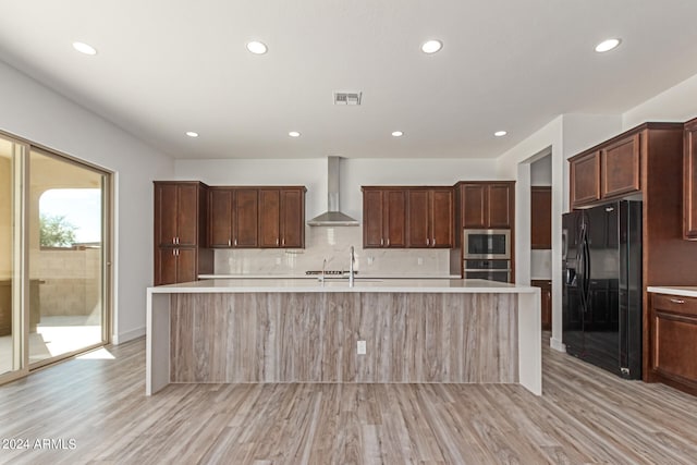 kitchen with light hardwood / wood-style floors, stainless steel appliances, wall chimney range hood, and an island with sink