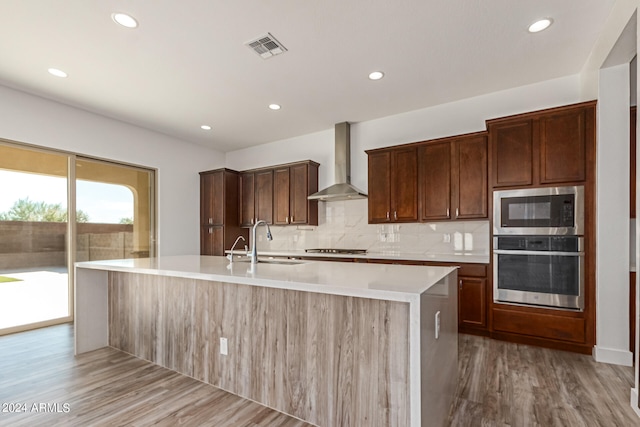 kitchen featuring a kitchen island with sink, wall chimney exhaust hood, sink, light wood-type flooring, and appliances with stainless steel finishes