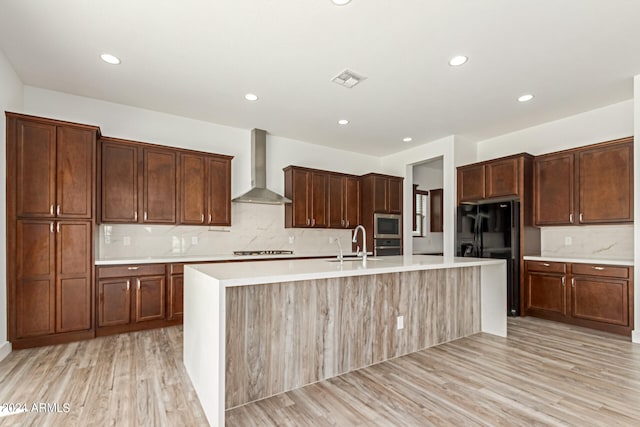 kitchen with decorative backsplash, wall chimney range hood, an island with sink, stainless steel appliances, and light wood-type flooring