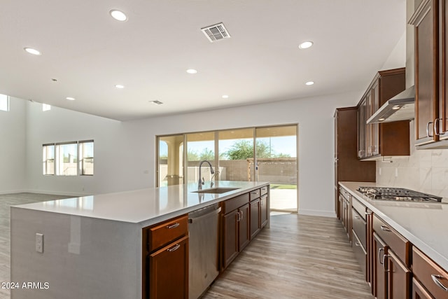 kitchen with wall chimney exhaust hood, light hardwood / wood-style flooring, an island with sink, sink, and appliances with stainless steel finishes