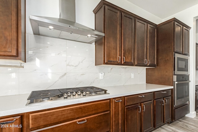 kitchen featuring tasteful backsplash, appliances with stainless steel finishes, dark brown cabinets, wall chimney exhaust hood, and light hardwood / wood-style flooring