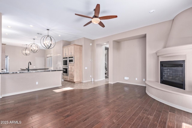 unfurnished living room featuring a large fireplace, ceiling fan with notable chandelier, a sink, and dark wood finished floors