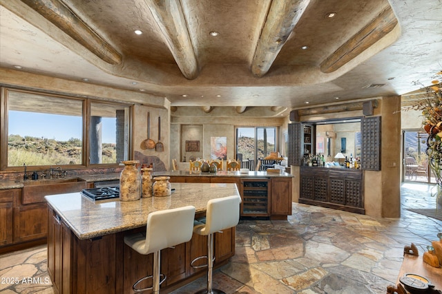 kitchen featuring beam ceiling, a kitchen island, light stone countertops, a kitchen bar, and beverage cooler