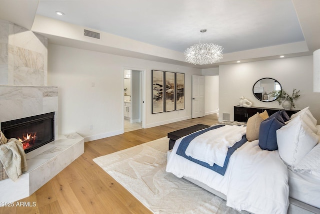 bedroom with connected bathroom, a tray ceiling, a high end fireplace, light hardwood / wood-style floors, and a chandelier