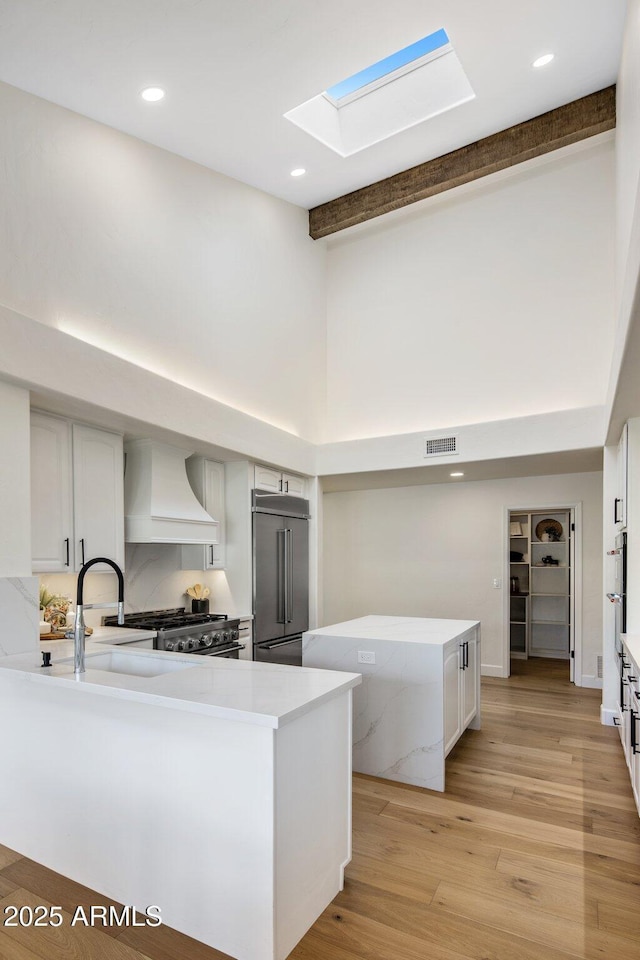 kitchen with a skylight, high quality appliances, white cabinets, custom range hood, and light hardwood / wood-style flooring