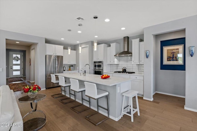 kitchen with stainless steel appliances, white cabinetry, decorative light fixtures, wall chimney range hood, and light wood-type flooring