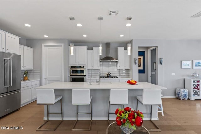 kitchen with a center island with sink, appliances with stainless steel finishes, wall chimney exhaust hood, decorative light fixtures, and white cabinets