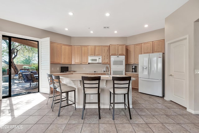 kitchen featuring light tile patterned floors, an island with sink, light brown cabinets, white appliances, and a kitchen breakfast bar