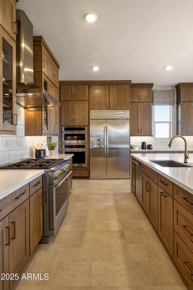 kitchen featuring stainless steel appliances, a sink, light countertops, brown cabinets, and wall chimney exhaust hood