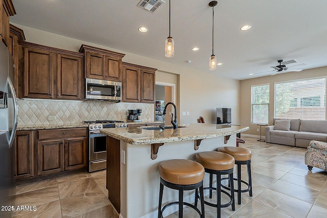 kitchen featuring appliances with stainless steel finishes, sink, hanging light fixtures, light stone countertops, and a center island with sink