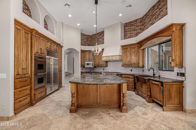 kitchen featuring a kitchen island with sink, dark stone countertops, a towering ceiling, hanging light fixtures, and built in appliances