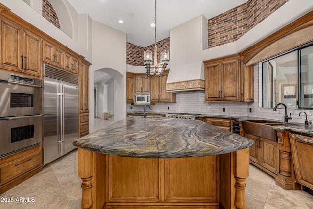 kitchen featuring a towering ceiling, an island with sink, built in appliances, and dark stone counters