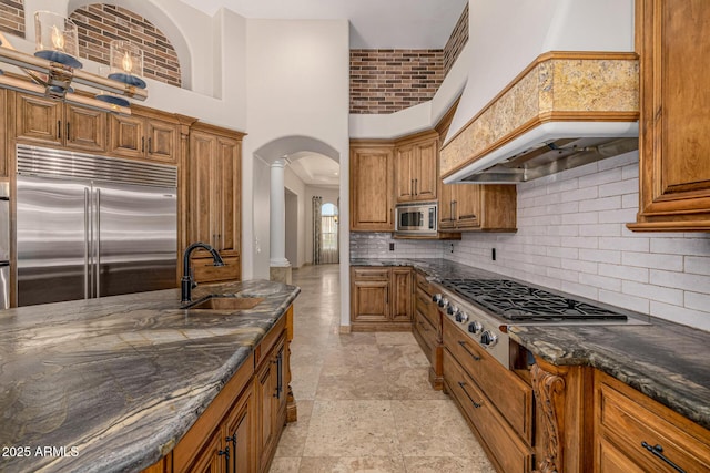 kitchen with a high ceiling, dark stone counters, built in appliances, decorative backsplash, and sink