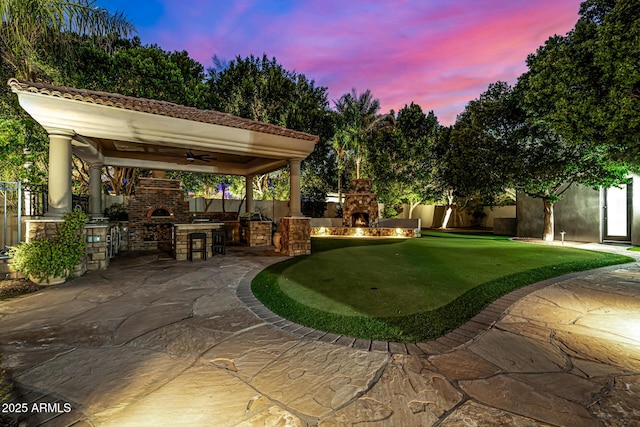 patio terrace at dusk with ceiling fan, a gazebo, and an outdoor brick fireplace