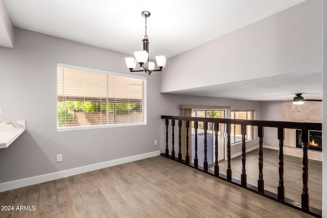 empty room with ceiling fan with notable chandelier, a warm lit fireplace, baseboards, and wood finished floors